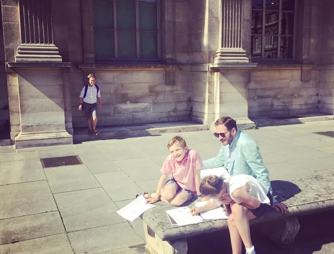 Père et enfants remplissant un carnet d'énigmes sur un banc du Louvre lors d'un jeu de piste en famille à Paris
